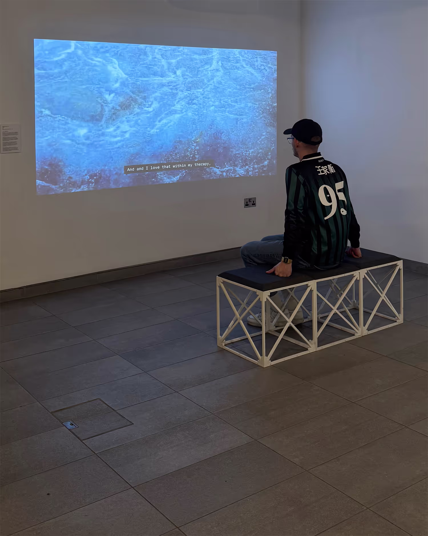 A man sits on a seat in an art gallery, watching a projected artwork play against the wall. Crashing waves can be seen in the video. 