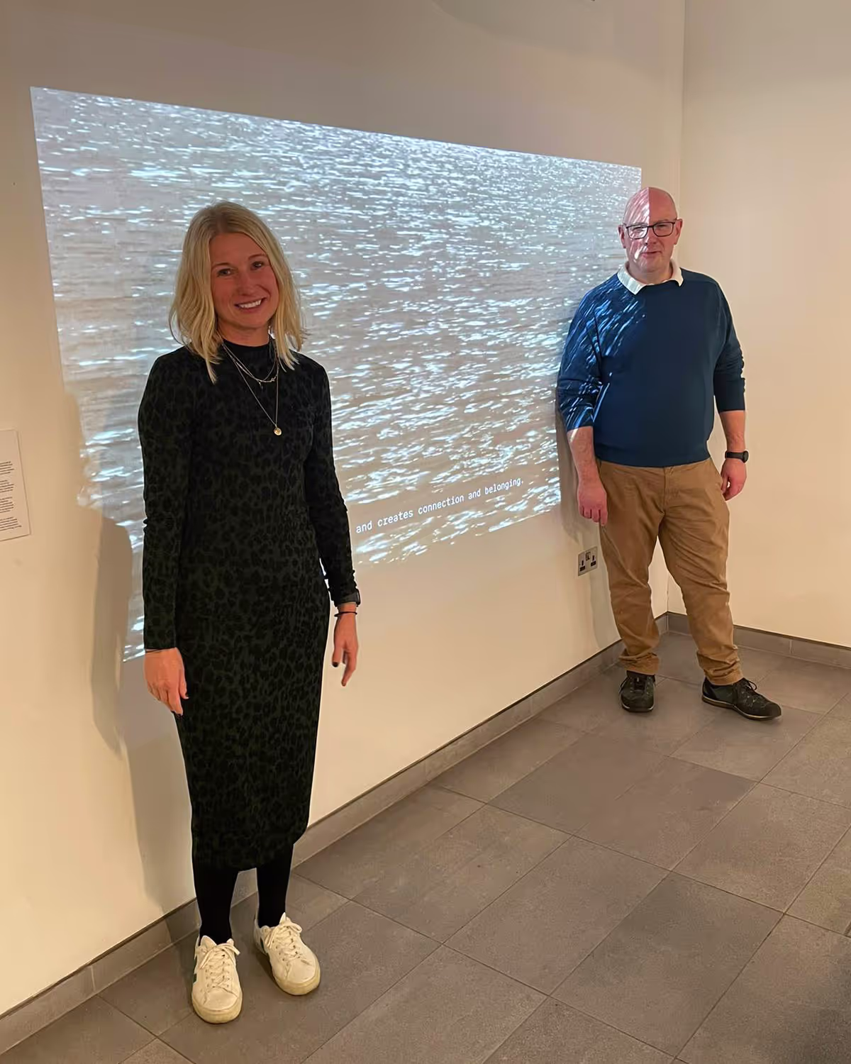 A man and a woman stand beside a projected video in a gallery space.