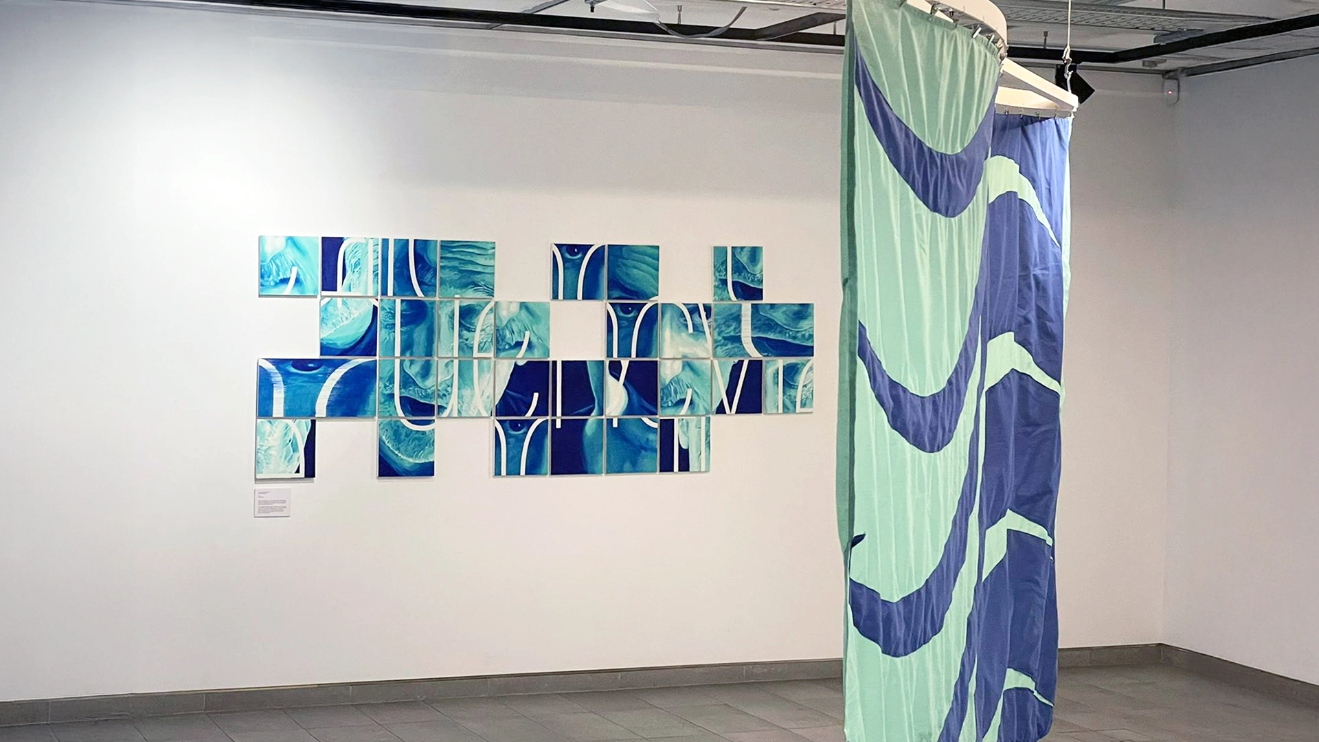 A blue and green painted artwork on a white wall. The artwork is made up of 25 squares, disorganised like a puzzle, showing portraits of two men speaking. A flag in the same colours hangs in the foreground. 